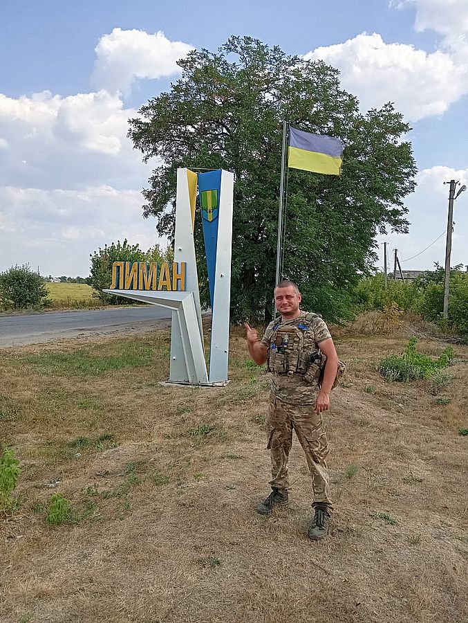 Український військовий у формі стоїть біля стели «Лиман» та державного прапора України. Фото зроблене на в’їзді до міста після бойових дій на Донеччині. Боєць показує жест перемоги на тлі українського прапора та літнього неба.