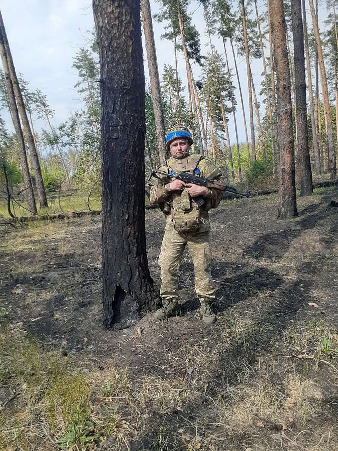 Український військовий зі зброєю стоїть у випаленому лісі серед пошкоджених сосен. На бійцеві військова форма, бронежилет і синя стрічка на шоломі. Фото передає атмосферу бойових дій та наслідки обстрілів у лісовій місцевості на фронті.