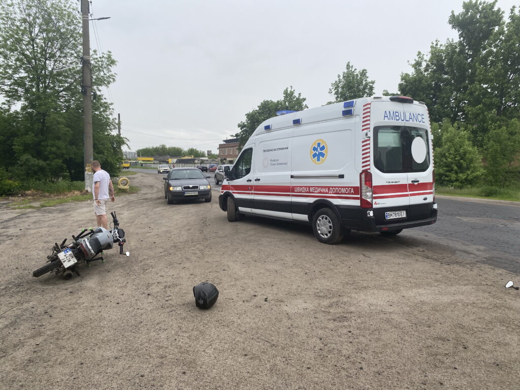Автомобіль швидкої допомоги на місці ДТП в Охтирці, де після зіткнення з легковиком перекинувся мотоцикл. На узбіччі лежать мотоцикл та мотошолом, обставини аварії з’ясовують правоохоронці.