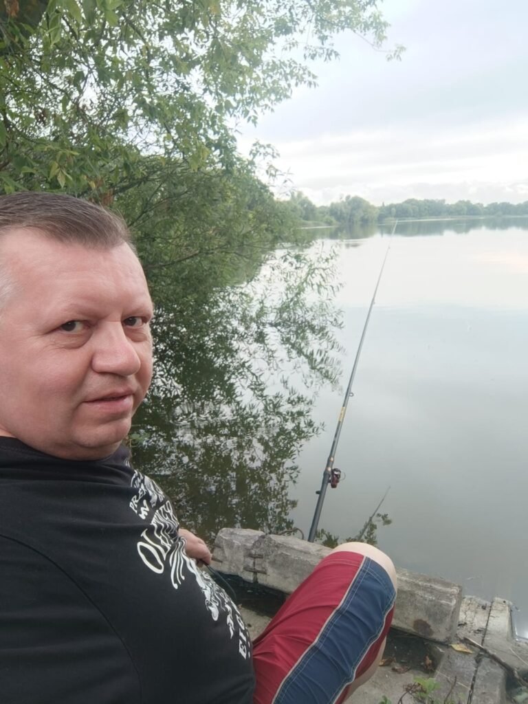 Віталій Кохан сидить із вудкою на березі спокійної водойми.